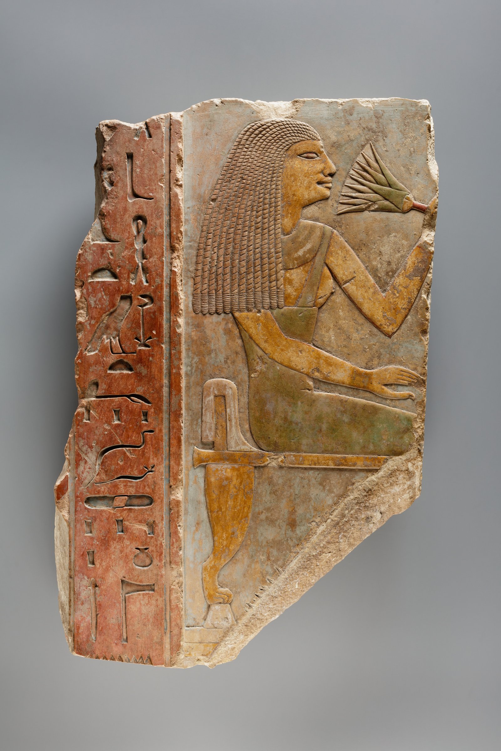 Painted limestone relief, Middle Kingdom Egypt: a seated woman with long hair lifts a water lily to her nose, a column of hieroglyphs on red at left