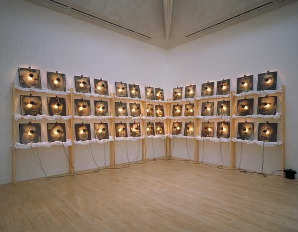 Christian Boltanski, The Reserve of Dead Swiss: ba hàng kệ gỗ xếp ảnh chân dung mờ, mỗi ảnh có một bóng đèn kẹp phía trên, dây điện buông xuống