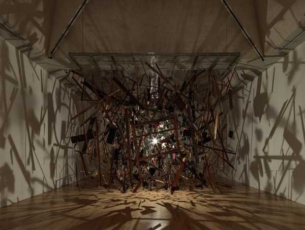 Cornelia Parker, Cold Dark Matter: An Exploded View: mảnh gỗ và đồ vật cháy xém treo lơ lửng quanh một bóng đèn, bóng đổ lên tường