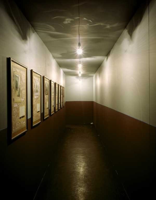 Ilya Kabakov, Labyrinth (My Mother's Album): hành lang gỗ hẹp, bóng đèn trần, khung ảnh và trang chữ treo dọc tường