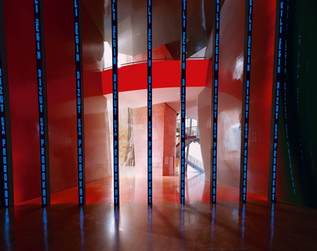 Jenny Holzer, Installation for Bilbao: chín cột đèn LED thẳng đứng chạy chữ sáng, dựng giữa không gian tường đỏ và kim loại cong của Guggenheim Bilbao
