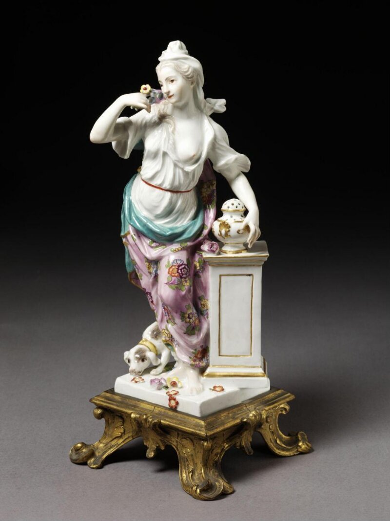 Tượng sứ Meissen Smell: phụ nữ mặc áo trắng và khăn choàng hồng hoa văn, đưa bó hoa lên mũi, tay đặt trên lư hương trên bệ vuông, con chó dưới chân, đế đồng mạ vàng