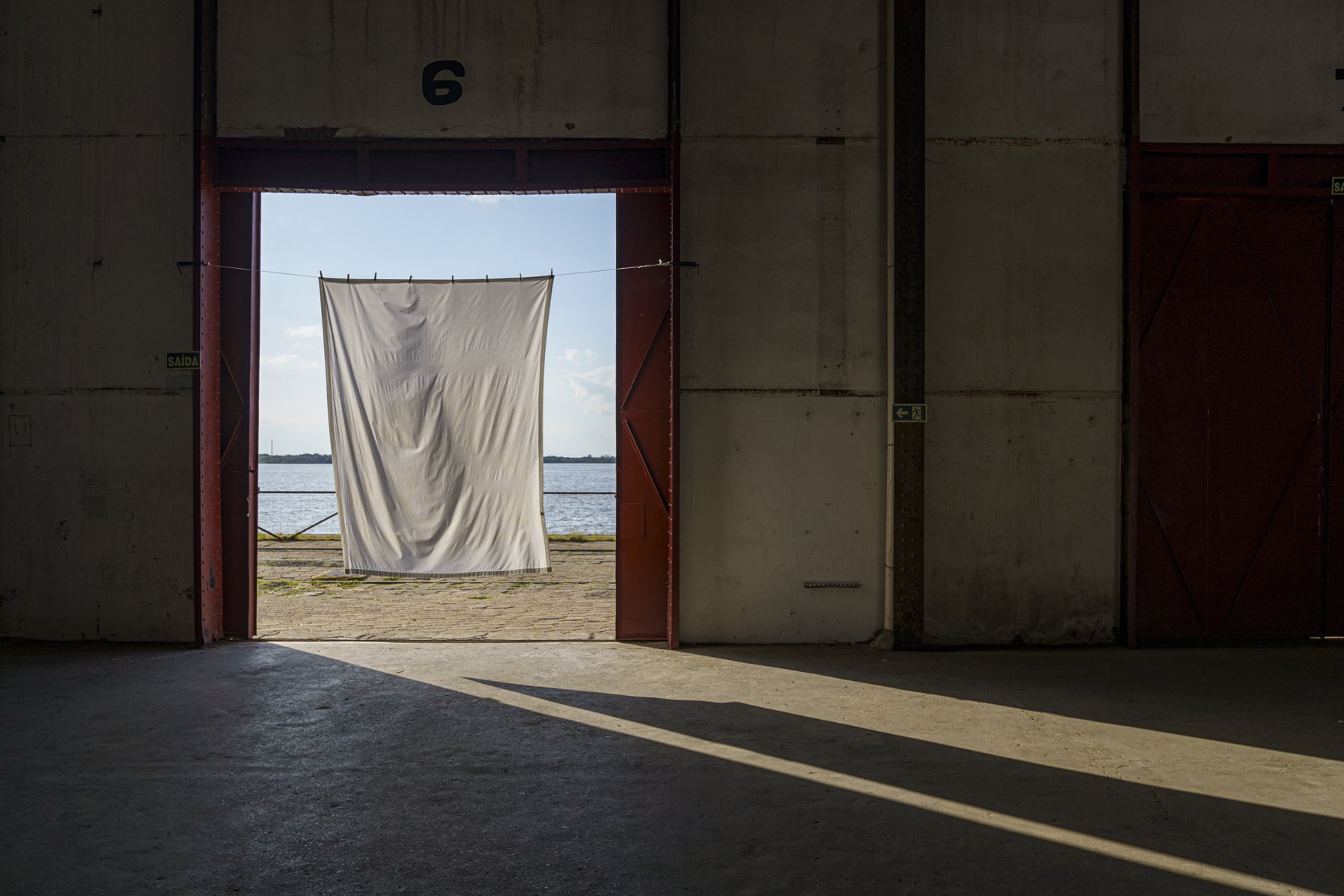 Lágrimas, Terra e Crisântemo by Karola Braga: a scented white sheet on a clothesline before the Guaíba River, Porto Alegre