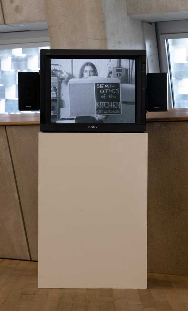 Martha Rosler, Semiotics of the Kitchen: người phụ nữ đứng sau bàn bếp, cầm dụng cụ nấu ăn, hình đen trắng