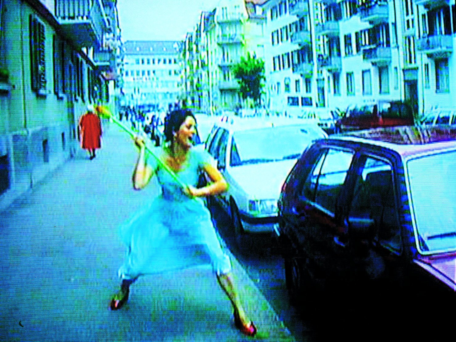 Pipilotti Rist, Ever Is Over All: phụ nữ váy xanh nhạt cầm cành hoa đi trên vỉa hè, cạnh hình chiếu những bông hoa đỏ