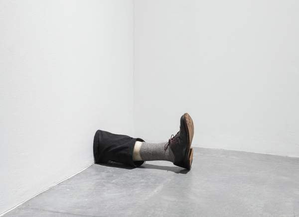 Robert Gober, Untitled: cẳng chân bằng sáp mặc quần sẫm màu, tất xám và giày da, thò ra từ chân tường ở góc phòng trắng